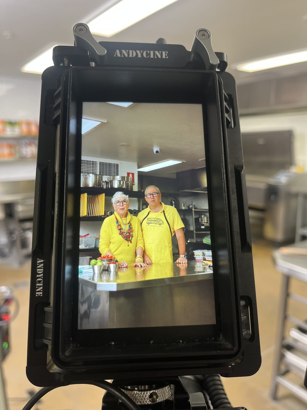 A camera monitor framing two OzHarvest volunteers in yellow aprons in the kitchen